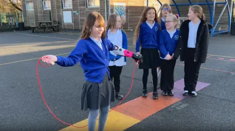 Zoey is in her blue school uniform and is smiling. She has brown hair and a fringe. Part of he bionic arm can be seen as the sleeve of her cardigan has been pushed up. The bionic arm has a white casing and black outlining. She is holding a pink skipping rope on a patterned plaground with five other children in the background