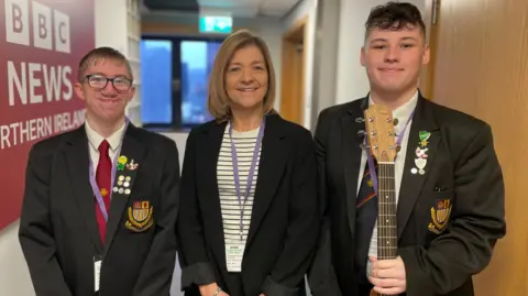 Music teacher Sharon Baird, wearing a black blazer over a white top with black stripes, stands between pupils Matthew McGibbon and Deacon Weary. Both teenage boys are dressed in shirt, tie and blazer, which has the school crest embroidered on the breast. Matthew is tall with dark hair and holds a guitar in his left hand. Deacon is shorter, with fair hair and glasses.