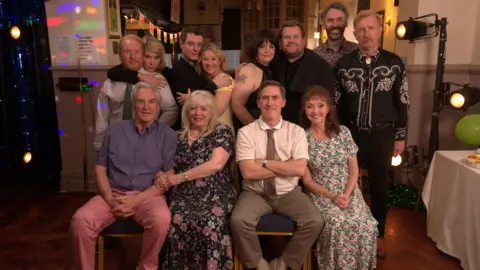 PA Images The main cast from Gavin and Stacey. Some of the cast members are sitting down on a chair and others are standing up. Everyone is smiling at the camera. 
They are on the set of the final episode from last year