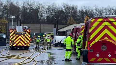 WMFS Six firefighters wearing brown uniforms and green high-visibility clothes. A fure engine is on the left and several other fire vehicles are in view as well as a white van. 