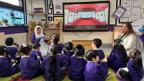 A class of nursery age children are sitting on the floor watching teachers demonstrate how to brush their teeth.  The children have their backs to the camera.  There is also a large screen on the wall playing a video about how to brush properly.