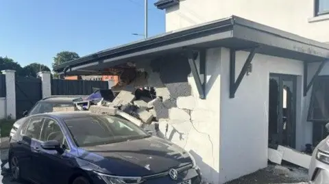 A white house with a navy Volkswagen Golf car parked outside. One wall of the house has been hit and is crumbling. 