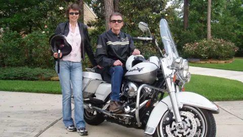 Chris and Denise Arthey A man sat on a motorbike and a woman stood next to the motorbike holding a helmet. They are posing for a photograph on a driveway. 