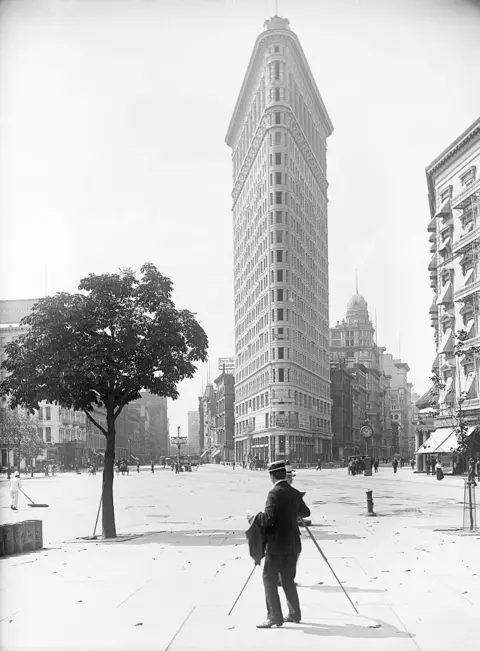 Corbis via Getty Images historic black and white photo showing a photographer in front of triangular Flatiron Building