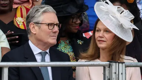 Getty Images Sir Keir Starmer and Victoria Starmer sit to watch the parade. He is wearing a dark suit with a white shirt and a grey tie, and she wears a pale pink dress with a white hat.