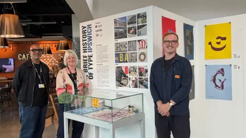 Supplied A man and a woman standing next to a display cabinet and a large white board on which there are close-up images of letters and a description about an exhibition. On the right side of the cabinet is a man. They are all smiling and looking into the camera. 