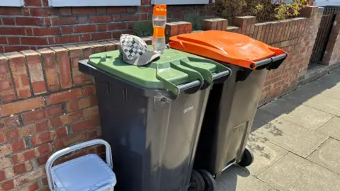 Two large outdoor wheelie bins placed on a pavement beside a low brick wall in front of a house. One bin has a green lid and the other has an orange lid.