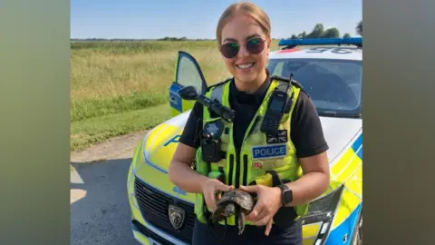 A female police officer is holding a tortoise and standing in front of a police car. She has fair hair that is pulled back and is wearing sunglasses. There are fields in the background.