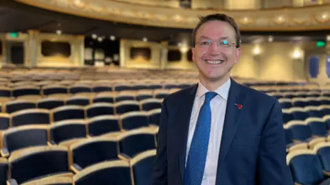 A man wearing a navy suit white shirt and light blue tie, standing in a theatre with rows of chairs behind him.