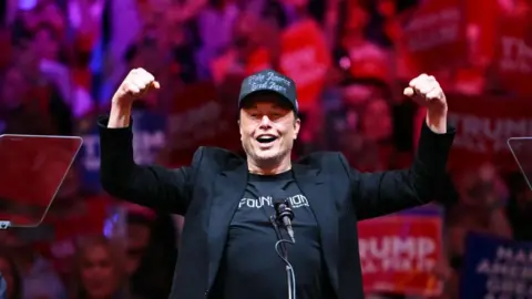 Getty Images Elon Musk, wearing black, holds his arms up victoriously at a rally for Donald Trump in October