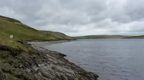Geograph/Richard Law Claerwen Reservoir seen from the shore surrounded by grassy hills and rocky sloping bank