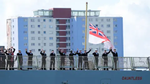 Royal Navy personnel wave from the deck of the Royal Navy Type 45 Destroyer HMS Dauntless as it sails from Portsmouth Naval Base.