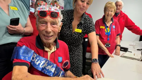 A man wearing a red top with a birthday banner has birthday glasses on his head, as colleagues look on behind him. 
