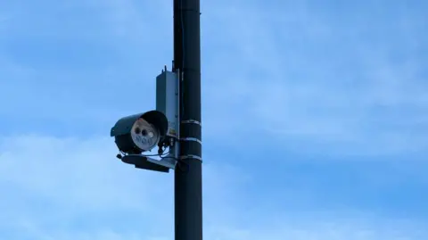 A picture of a black ANPR camera on a black pole. The sky is blue in the background.