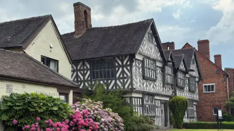 A black and white, three-storey Elizabethan building is positioned alongside a more modern brick-built building which is painted cream. There is a pink rhododendron bush in front of the brick building as well as a small lawned area.