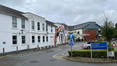 The side of entrance of a hospital in Guernsey. There is a hedge with a blue sign in front of it. One car is parked in a spaces immediately behind the hedge. One of the buildings is white with a rainbow painted across the side.
