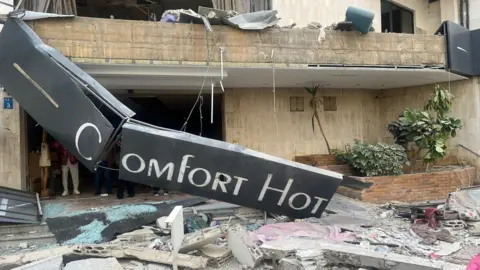 Alice Cuddy/BBC A broken sign reading Comfort Hotel hangs outside the front of a badly damaged building with debris on the pavement