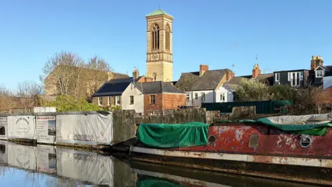 Jericho Wharf. It is right on the Oxford Canal, with a derelict canal boat sitting next to developers panelling. In the background is a church tower.