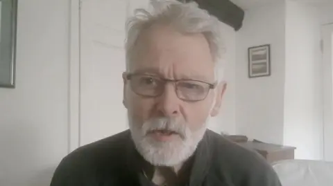 A man wearing a grey zip up jumper and glasses. He has a white beard and white hair. Behind him is a room. The walls are white and there are photo frames and art work on the wall. There appears to be a sofa to the right.