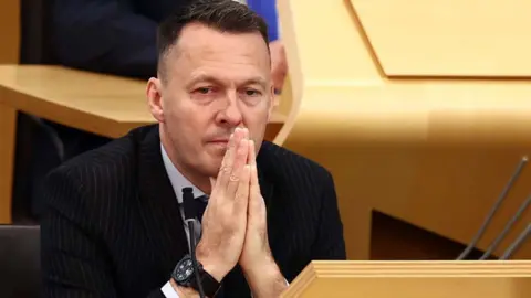 Russell Findlay looking sitting in the Scottish Parliament chamber with his hands together in front of him