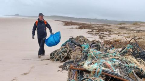 A man walks past a huge pile of fishing waste consisting of ropes and pots on a wide empty beach 