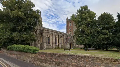 Google External view of St Peter's Church with its brick built tower. A number of large mature trees can be seen in the churchyard. The edge of the church has a brick wall, which drops down to St Peters Road in the foreground. 