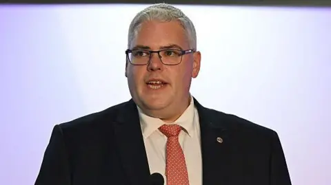 Getty Images Gavin Robinson speaking. He is wearing a black suit blazer, white shirt and a pink/orange tie. He is wearing black framed glasses and has grey hair.