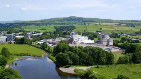 White factory in a green landscape with a pond and several trees.