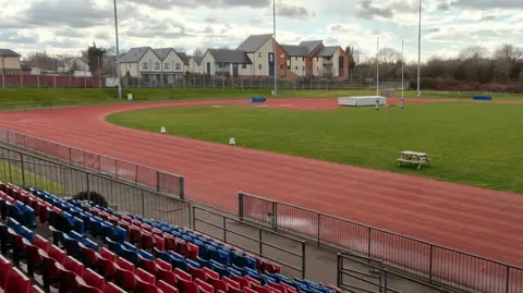 A red athletics track curving around the corner of an outdoor stadium. There are blue and red plastic seats in the foreground, and a rugby post in the distance.