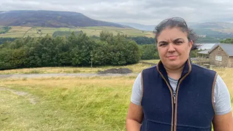 BBC A woman in a light blue t-shirt and navy bodywarmer. She is stood on a green field in a valley. At the other side of the valley is a moor, half of which has been badly burned. There is also a reservoir in the distance.