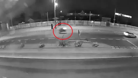 GYBC Black and white noise detection camera footage of Great Yarmouth seafront.  red circle highlights a motorbike with its lights on pulling a wheelie while travelling along the road 