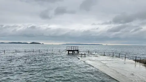 BBC A diving board on the edge of a Victorian open air swimming pool by he sea. Iron railings surround it.