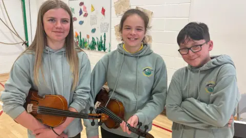Three young children in green hoodies, two with violins in the rest position.
