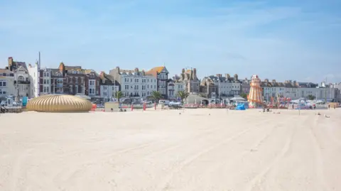 White sandy beach with attractions along its edge and buildings stretching behind.  It is a bright, sunny day.