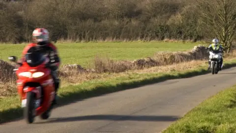 Two motorcyclists going along a country road. The bike closest to the picture is red. Around them are trees and fields.
