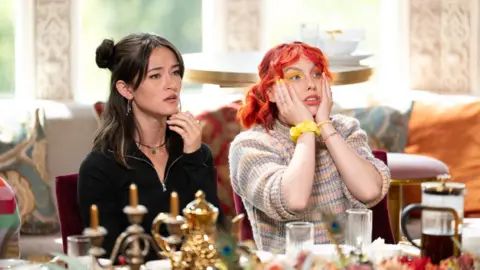 PA Media Jessie is shocked and has her hands over her face. She has bright orange hair. She is sat beside a fellow contestant who has dark brown hair and wearing a black hoodie. They are sat at a breakfast table. 