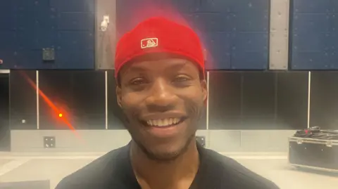 Actor Sean-Marco Msipha wearing a red baseball cap, smiling for the camera at a production space at the Aviva Studios building.
