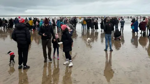 Peter Sawyer Hundreds of people wearing coats and warm weather outfits standing on a beach as people in the distance head into the sea.