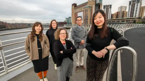 Northumbria University Jenny Alderson and Hayley Duff of Baltic; and Dr Ayse Ozbil Torun, Dr Bing Zhai and Dr Jiayi Jin of Northumbria University stand on a bridge across the Tyne River in front of the Baltic building.