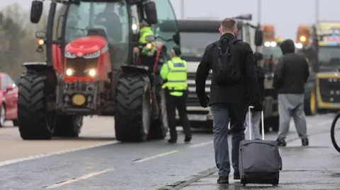 PA Media A man is pulling a suitcase along the side of a road. A number of tractors and trucks can bee seen on the road. Police are speaking to the person in the tractor at the head of the convoy.