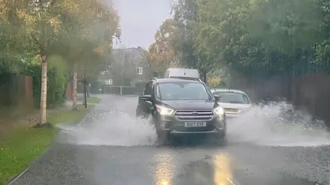 A car drives past a parked vehicle on a flooded suburban street lined with trees on both sides.