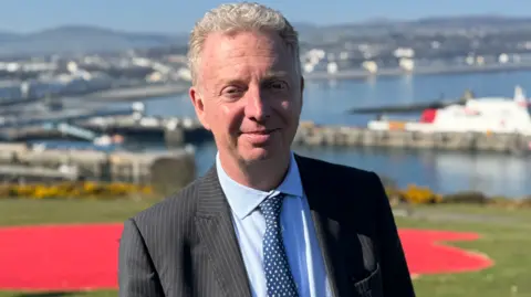 BBC Alex Allinson is wearing a suit, with a black pinstripe jacket, a light blue shirt, and a dark blue spotted tie. He has grey, short, wavy hair and is standing outside on a sunny day, with Douglas Bay behind him.