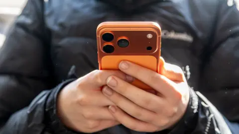 A close up shot of a 15 year old's hands holding a smartphone with an orange case, taken in Bath in January.