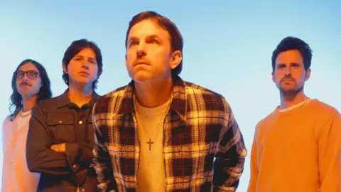 The four band members of Kings of Leon staring into the camera against a blue background. From left to right: Nathan Followill, who has long brown hair and a moustache, Matthew Followill, in a denim shirt, with his arms crossed. Cale Followil, leaning forward, with short brown hair and a crucifix around his neck, a checked shirt and a white top underneath. To his right, Jared Followill whose brown hair is swept back; he is wearing a yellow jumper. 