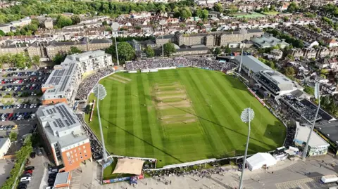 PA Media An aerial shot of the County Ground in Bristol during a T20 cricket match. Temporary stands full of spectators are visible, as are the high-rise flats that overlook the stadium. It is a sunny evening with the floodlights casting shadows across the pitch. The image is taken from a drone