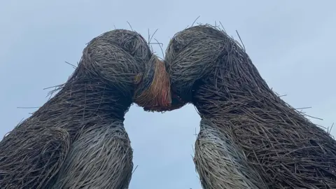 A side on shot of the two puffin sculptures with their heads together. The willow is frayed and coming off in many places and the paintwork is faded. A grey-ish sky is visible in the background.