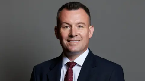 UK Parliament Stuart Anderson has short brown hair. He is wearing a dark suit jacket with a white shirt and red tie with gold spots on it