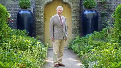 Charles III stands on a garden path leading to a yellow gate in a brick wall. There is well-kept foliage on either side of him and he is wearing a beige blazer, trousers and brown shoes.