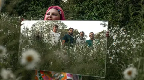 Biskee Brisht Lead vocalist Ruby Biscoe-Taylor stands in a field holding a mirror to which the rest of the band members can be seen.