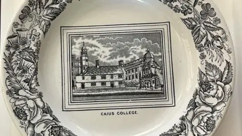 A white plate is decorated with a view of Caius Court in its centre, in black, and the words "Caius College" printed beneath. Around the edges are black drawings of flowers and foliage
